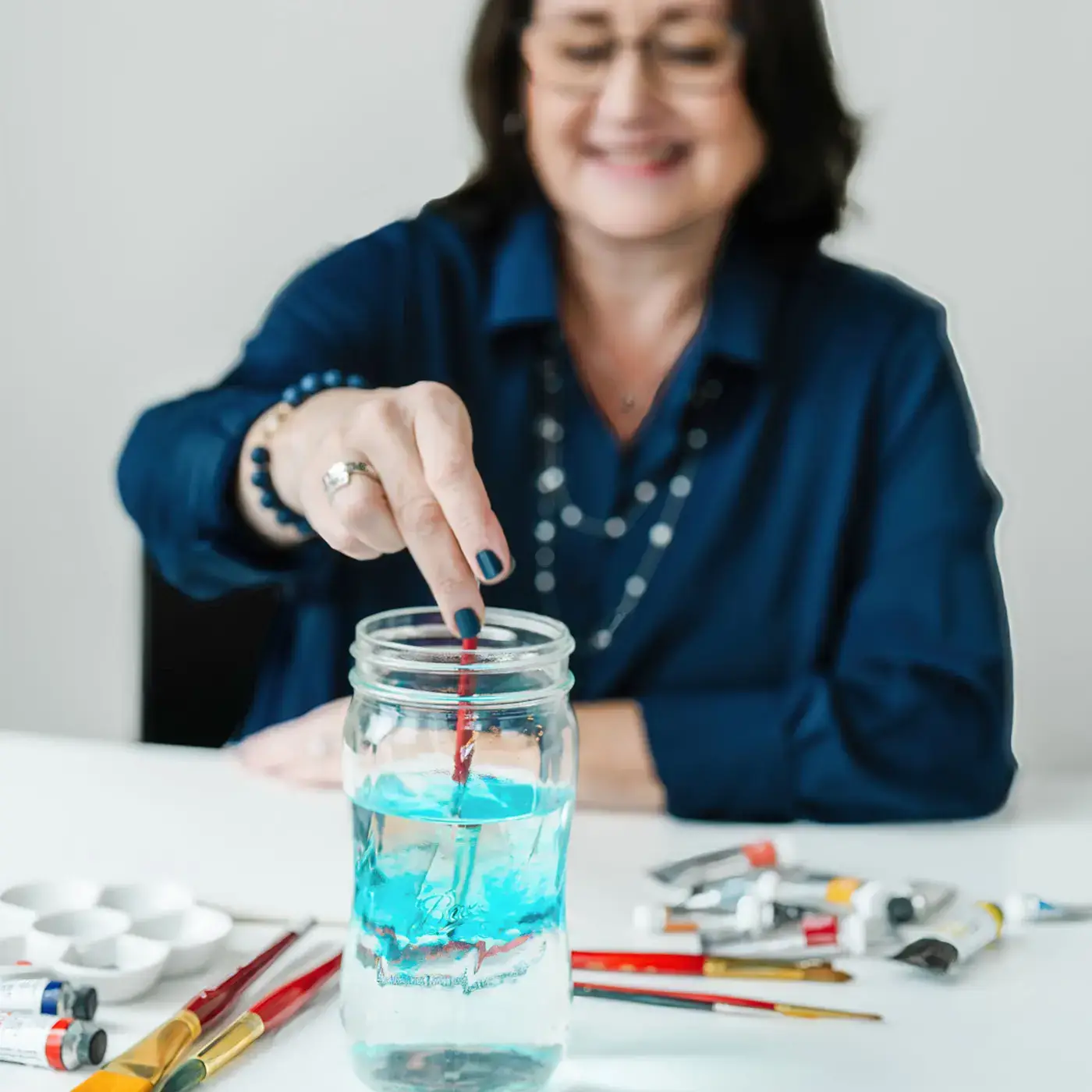 Artist stirring blue watercolor paint in a glass jar of water at a work table, with paintbrushes and tubes of paint nearby.