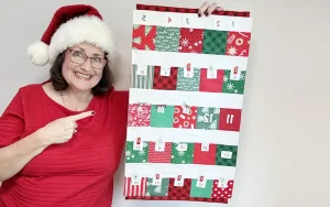 Tara Reed in a Santa hat holding a handmade fabric Advent calendar with 25 pockets. Beginner-friendly Christmas sewing pattern made from fabric scraps with printable numbers and video tutorial.