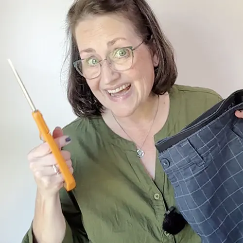 Cutting Apart Men’s Shorts: Smiling sewing instructor holding orange-handled scissors and a pair of dark plaid men’s shorts, demonstrating how to reuse clothing for sewing projects.